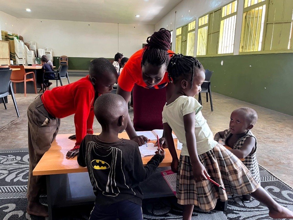 A KPDDI volunteer helps children with their reading at a table in the community hall